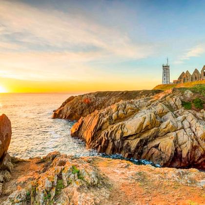 Red and white striped lighthouse in Plougonvelin on rocky cliffs at sunset, with ruins and calm ocean under golden sky.