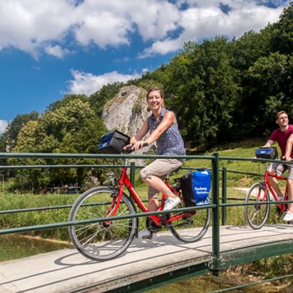 Zwei Radfahrer mit Packtaschen überqueren eine Holzbrücke mit grünem Geländer. Dichter Wald und Felsklippe im Hintergrund unter blauem Himmel.