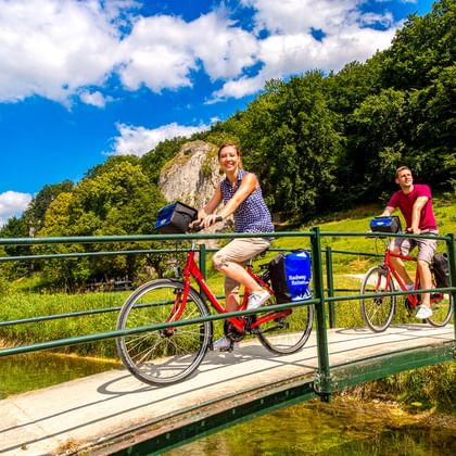 Zwei Radfahrer auf einer Metallbrücke über einen Bach bei Schelklingen an der Donau. Grüne Hügel und Felsen unter blauem Himmel.