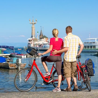 Couple with bicycles viewing Konstanz harbor on Lake Constance. Ferry boat and Imperia statue visible, boats moored in sunny harbor.