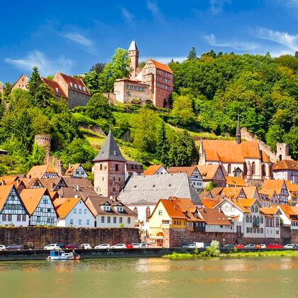Historische Stadt Hirschhorn am Neckar mit Fachwerkhäusern, orangen Dächern und einer Burg auf dem Hügel unter blauem Himmel.