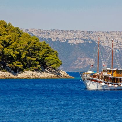 Traditionelles Holzsegelschiff MS Orkan auf ruhigem blauen Wasser, mit einer grünen bewaldeten Insel und felsigen Bergen im Hintergrund.