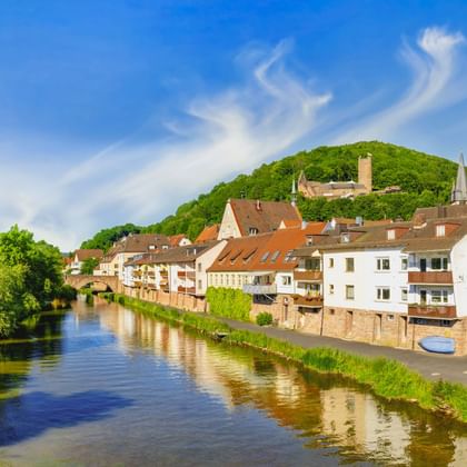 Historischer Hexenturm am Fluss in Gemünden mit traditionellen Häusern am Ufer und der Scherenburg im Hintergrund.