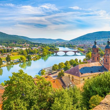 Luftaufnahme von Miltenberg mit rotgedeckten Gebäuden, Kirche mit zwei Türmen, Main mit Steinbrücke und grünen Hügeln unter blauem Himmel.
