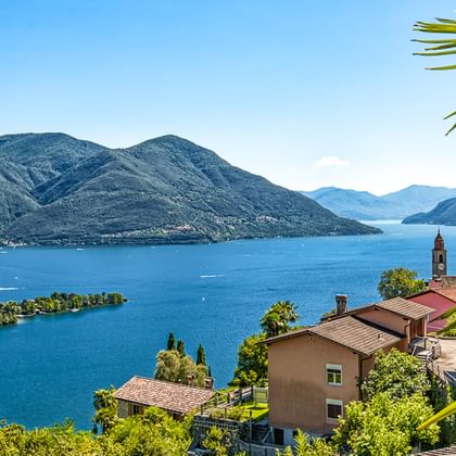 Malerischer Blick auf den Lago Maggiore mit blauem Wasser, kleinen Inseln und Bergen. Häuser mit roten Dächern und Kirchturm im Vordergrund, Palmwedel sichtbar.