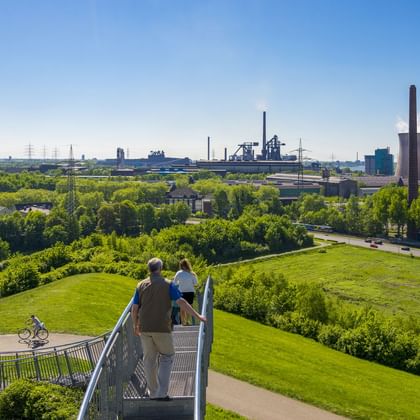 Erhöhter Gehweg im Landschaftspark Duisburg-Nord mit Besuchern, Blick auf grüne Landschaft, Radfahrer auf Weg und Industriebauten im Hintergrund.