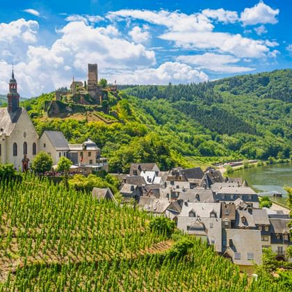 Dorf Beilstein mit Kirche und Burgruine Metternich auf Hügel, umgeben von Weinbergen und bewaldeten Hügeln entlang der Mosel.
