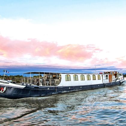 Weiß-blaues Flussschiff L'Estello mit Fahrrädern an Deck auf ruhigem Wasser in der Provence unter rosa-blauem Abendhimmel.