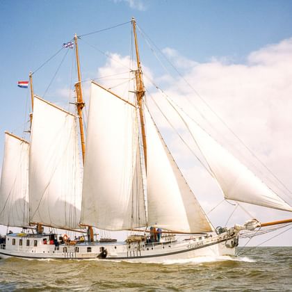 Dreimastklipper Elizabeth segelt mit weißen Segeln vor blauem Himmel mit Wolken. Das weiße Schiff bewegt sich durch ruhiges Wasser.