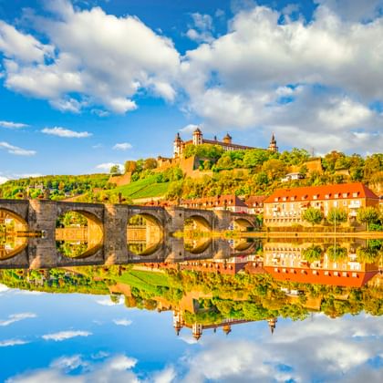 Festung Marienberg auf grünem Hügel über Würzburg mit Steinbrücke und bunten Häusern, die sich im Main unter blauem Himmel spiegeln.