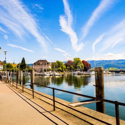 Lindauer Hafenpromenade mit historischem Leuchtturm, vertäuten Segelbooten und Löwenstatue am Hafeneingang unter blauem Himmel mit Bergen im Hintergrund.