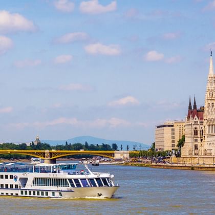 Weißes Flusskreuzfahrtschiff auf der Donau mit dem ungarischen Parlamentsgebäude und roter Kuppel am Flussufer in Budapest.