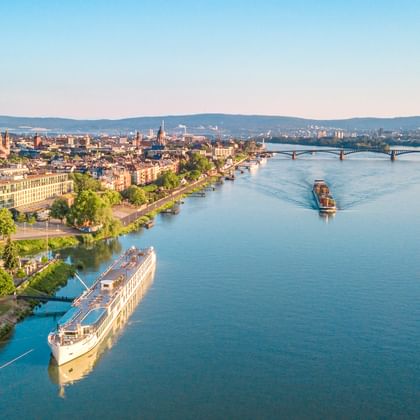 Luftaufnahme eines Flusskreuzfahrtschiffs auf dem Main in Mainz mit Stadtpanorama, Brücken und grünen Ufern unter klarem Himmel.