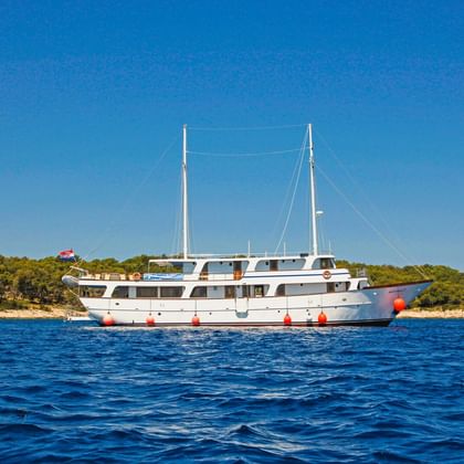 Weiße Motoryacht MS Azimut mit zwei Masten vor Anker auf ruhigem blauen Wasser. Grüne bewaldete Küstenlinie im Hintergrund unter klarem Himmel.
