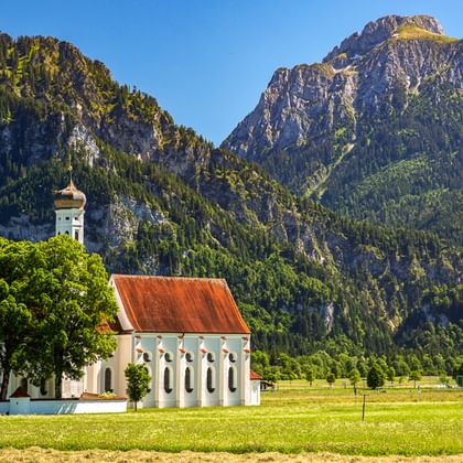 Weiße Kapelle mit rotem Dach auf grüner Wiese, umgeben von bewaldeten Bergen. Schloss Neuschwanstein sichtbar auf entferntem Hang unter blauem Himmel.