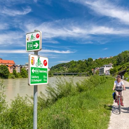 Zwei Radfahrer auf einem asphaltierten Weg entlang eines grünen Flusses. Radwegschilder weisen zum Tauernradweg und Salzburg. Historische Stadt mit Kirchturm sichtbar.
