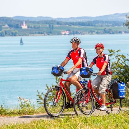 Zwei Radfahrer in roten Trikots mit Tourenrädern pausieren auf einem Weg mit Blick auf den Bodensee bei Dingelsdorf, mit Segelbooten und fernem Ufer.