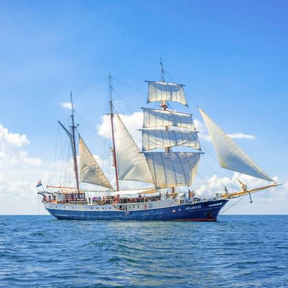 Dreimastiger Großsegler Atlantis mit weißen Segeln auf ruhigem blauen Meer unter teilweise bewölktem Himmel.