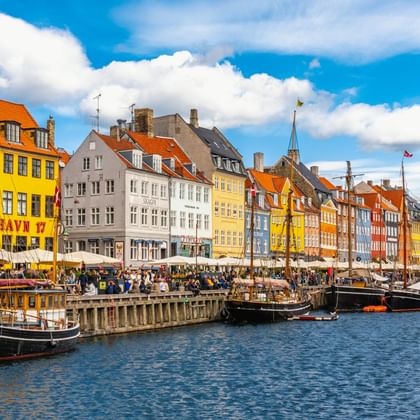 Historische Nyhavn-Uferpromenade in Kopenhagen mit bunten Stadthäusern, traditionellen Holzbooten im Kanal und Besuchern unter blauem Himmel.