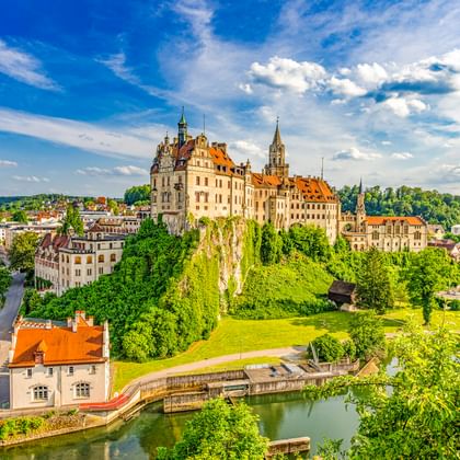 Hohenzollernschloss in Sigmaringen auf einem grünen Hügel über der Donau, umgeben von Bäumen und historischen Gebäuden unter blauem Himmel.