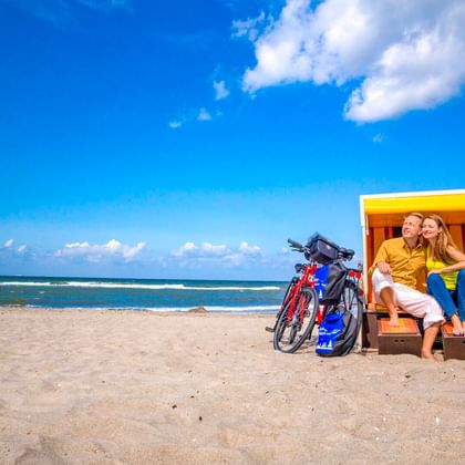 Couple relaxing in a yellow-white beach chair with two bicycles parked beside them on a sandy beach in Wustrow under a blue sky.