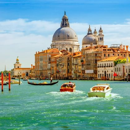 Boats and gondola on Venice's Canale Grande with the iconic dome of Santa Maria della Salute basilica and historic buildings under blue sky.