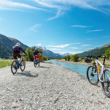 Zwei Radfahrer auf einem Schotterweg entlang des türkisfarbenen Inn-Flusses, mit einem geparkten Fahrrad im Vordergrund und Alpenbergen unter blauem Himmel.