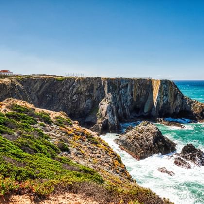 Leuchtturm mit rotem Dach auf dramatischen Felsenklippen an Portugals Atlantikküste. Türkisfarbene Wellen brechen an dunklen Felsen unter grüner Vegetation.