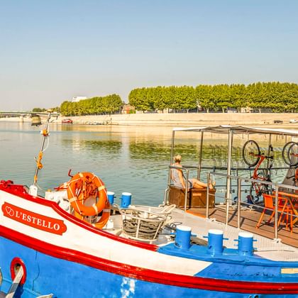 L'Estello Boot mit blauem und rotem Rumpf am Fluss in Frankreich. Orange Fahrräder auf Deck mit Fahrradabstellbereich unter Überdachung.