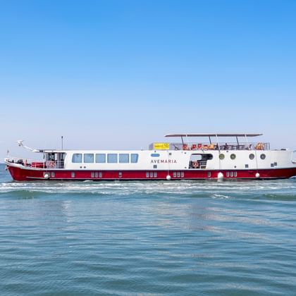 MS Ave Maria Flusskreuzfahrtschiff mit rot-weißem Rumpf auf ruhigem blauen Wasser unter klarem Himmel. Das Schiff hat mehrere Decks und Bullaugen.
