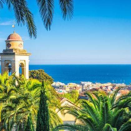 Weißer Glockenturm mit Kuppel, umrahmt von Palmen, mit Blick auf eine Küstenstadt an der italienischen Riviera und blauem Mittelmeer.