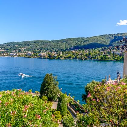 Klassische Statue in einem blühenden Garten mit Blick auf den Lago Maggiore mit grünen Hügeln und blauem Wasser. Ein Boot ist auf dem See sichtbar.