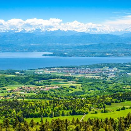 Panoramablick vom Jura Vaudois mit grünen Wäldern und Feldern, einem großen blauen See und schneebedeckten Alpen unter blauem Himmel mit Wolken.