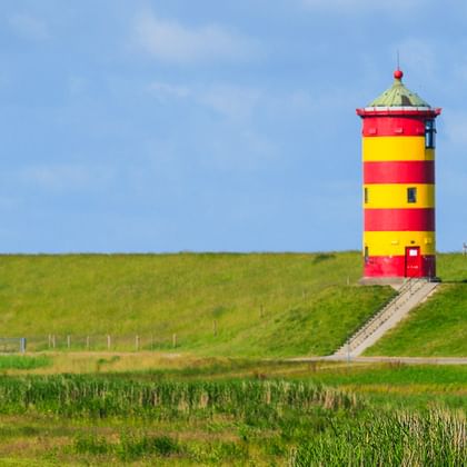 Rot-gelb gestreifter Leuchtturm auf grünem Deich mit Weg. Marschland im Vordergrund, blauer Himmel darüber.