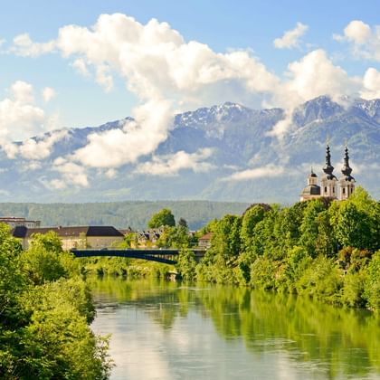 View of Villach along the Drau River with green vegetation, a church with twin spires, and snow-capped mountains under a cloudy sky.