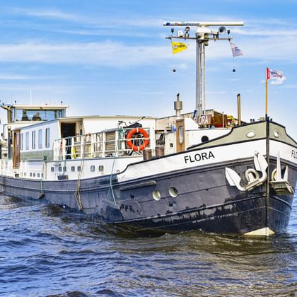 MS Flora, ein weiß-schwarzes Fahrgastschiff mit blauen Fenstern, fährt auf ruhigem Wasser unter blauem Himmel mit weißen Wolken.