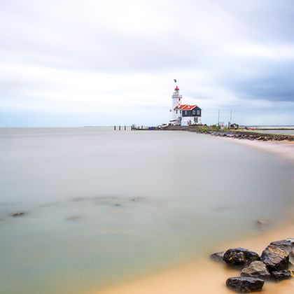 Weißer Leuchtturm mit rotem Dach auf einem Steg am IJsselmeer. Sandstrand mit Steinen im Vordergrund unter bewölktem Himmel.