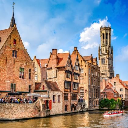 Mittelalterliche Backsteingebäude säumen einen Kanal in Brügge mit dem Belfried im Hintergrund. Ein Ausflugsboot fährt vorbei.