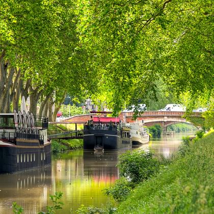 Zwei Radfahrer auf einem von Bäumen gesäumten Treidelpfad am Canal du Midi mit vertäuten Booten und einer Brücke im Hintergrund.