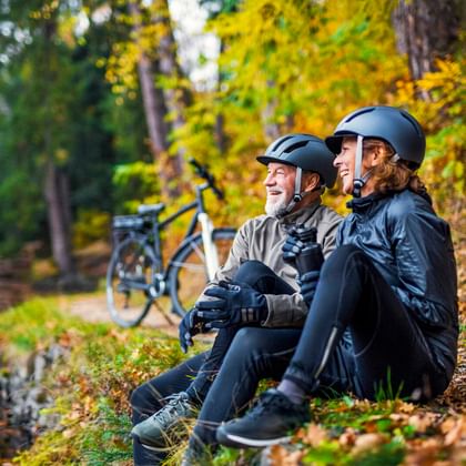 Zwei Radfahrer mit Helmen sitzen an einem Waldbach im Herbst, E-Bikes hinter ihnen geparkt. Buntes Herbstlaub umgibt die Szene.
