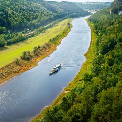 Luftaufnahme der Elbe bei Rathen, die sich durch grünes Tal mit bewaldeten Klippen schlängelt. Ein Raddampfer fährt auf dem ruhigen Wasser.