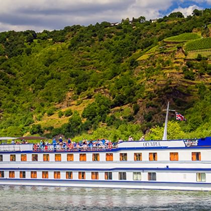 Weißes Flusskreuzfahrtschiff MS Olympia mit blauem Zierstreifen fährt auf ruhigem Fluss, mit grünen weinbewachsenen Hügeln im Hintergrund.