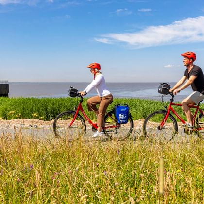 Zwei Radfahrer mit roten Helmen und blauen Taschen auf roten Rädern auf einem grasbewachsenen Deichweg in Brunsbüttel, mit Leuchtturm und Wasser.