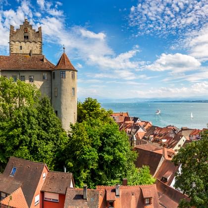 Mittelalterliche Burg Meersburg mit Rundtürmen über rotgedeckten Häusern, mit Blick auf den Bodensee mit Segelbooten unter blauem Himmel.