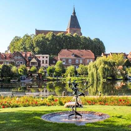 Blick über den Mölln Stadtsee mit Brunnen im Vordergrund, bunten Blumenbeeten, historischen Häusern am Ufer und Kirche auf dem Hügel.