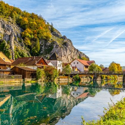 Essing village along the Altmühl river with traditional houses, wooden footbridge, and dramatic limestone cliffs in autumn colors.