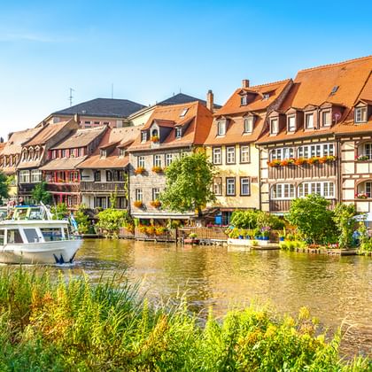 Colorful half-timbered houses with orange roofs along the Main River. A tour boat is moored at the waterfront, with green vegetation in foreground.