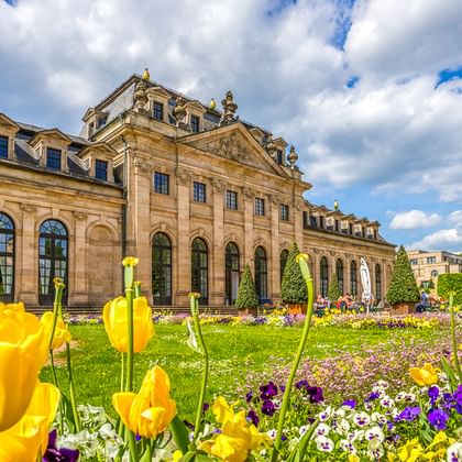 Baroque palace with ornate facade and blooming garden featuring yellow tulips and purple flowers under a cloudy blue sky.