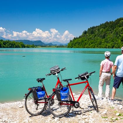 Zwei Radfahrer stehen am Ufer des türkisfarbenen Feistritzer Stausees mit zwei roten Tourenrädern, bewaldete Hügel und Berge im Hintergrund.