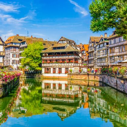 Traditionelle Fachwerkhäuser spiegeln sich in einem Kanal in Straßburg. Bunte Blumen säumen die Steinmauer unter blauem Himmel.
